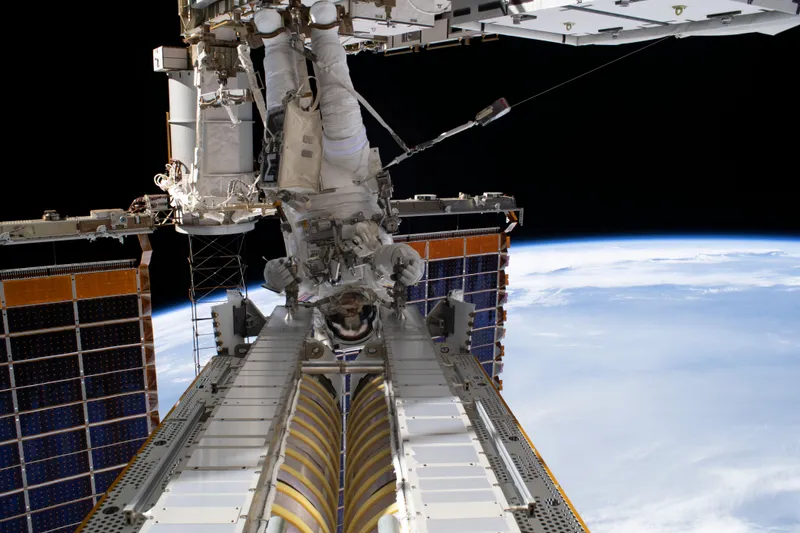 Seen from the space station's truss, an astronaut in a white spacesuit hangs upside down from the end of a robotic arm, gripping a long rolled-up solar array in a silver and gold frame that stretches towards the camera, with dark blue solar panels on either side and the white clouds of Earth's curved edge against black space