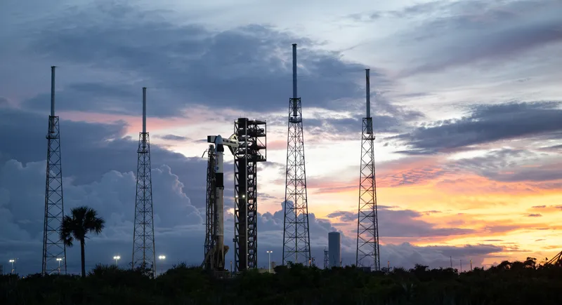 A Falcon 9 rocket with a Dragon capsule on top stands beside its launch tower at dusk, between four tall lightning masts, under a cloudy sky that turns orange on the right; a palm tree and dark scrub fill the foreground