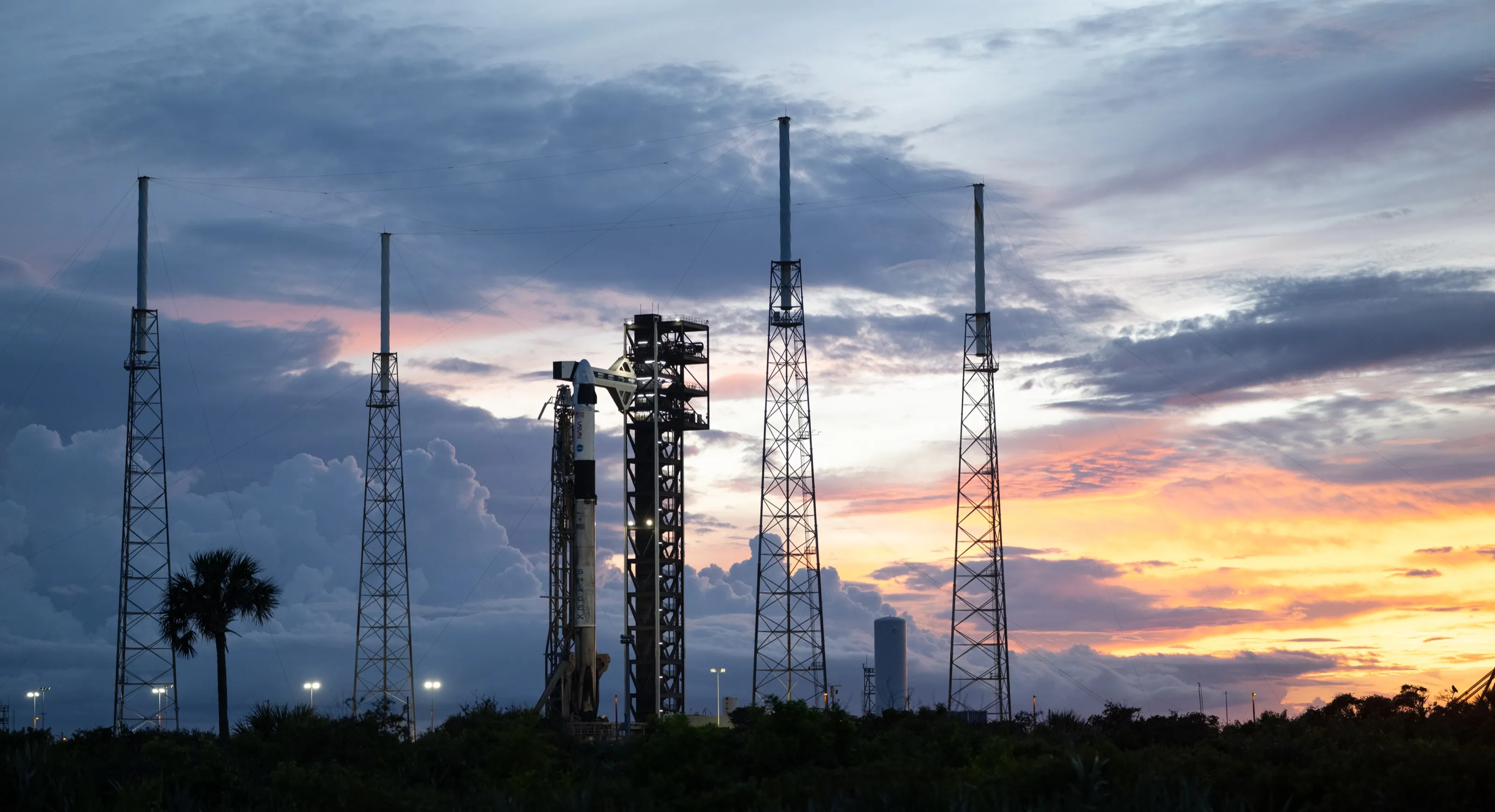 A Falcon 9 rocket with a Dragon capsule on top stands beside its launch tower at dusk, between four tall lightning masts, under a cloudy sky that turns orange on the right; a palm tree and dark scrub fill the foreground