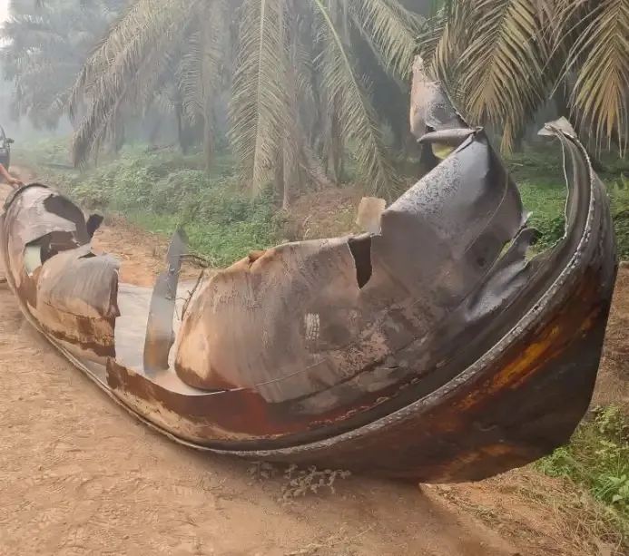 A large curved shell of torn, scorched metal, rust-brown and grey, lies on its side on a red dirt track between oil palms in a morning haze; jagged flaps of its skin stand up along the top, a hand rests on its left end, and a dark vehicle waits in the haze behind