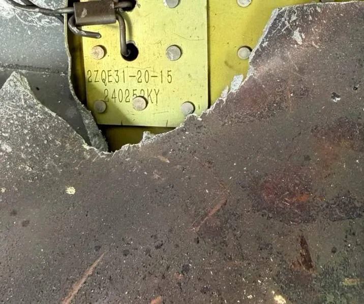 A close-up of a yellow-green metal plate held by rivets and a twisted wire, stencilled 2ZQE31-20-15 with a second code below it, partly covered by a torn grey sheet of metal with scorch and rust marks