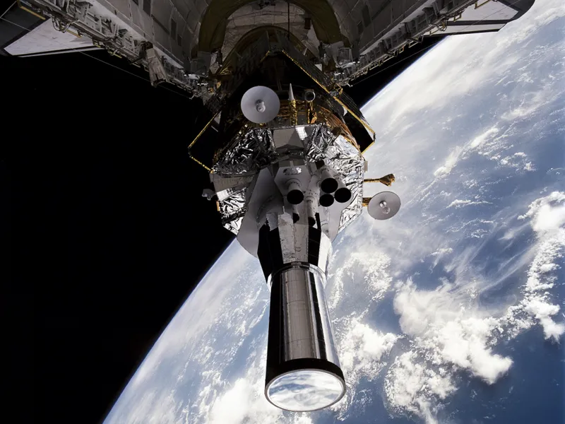 Seen from inside Atlantis's open cargo bay, a DSP satellite stands tilted up on its booster: gold and silver foil, two small dish antennas on its sides, a cluster of four nozzles, and a long, shiny tube pointing out over a blue Earth streaked with white cloud, the black of space at the left