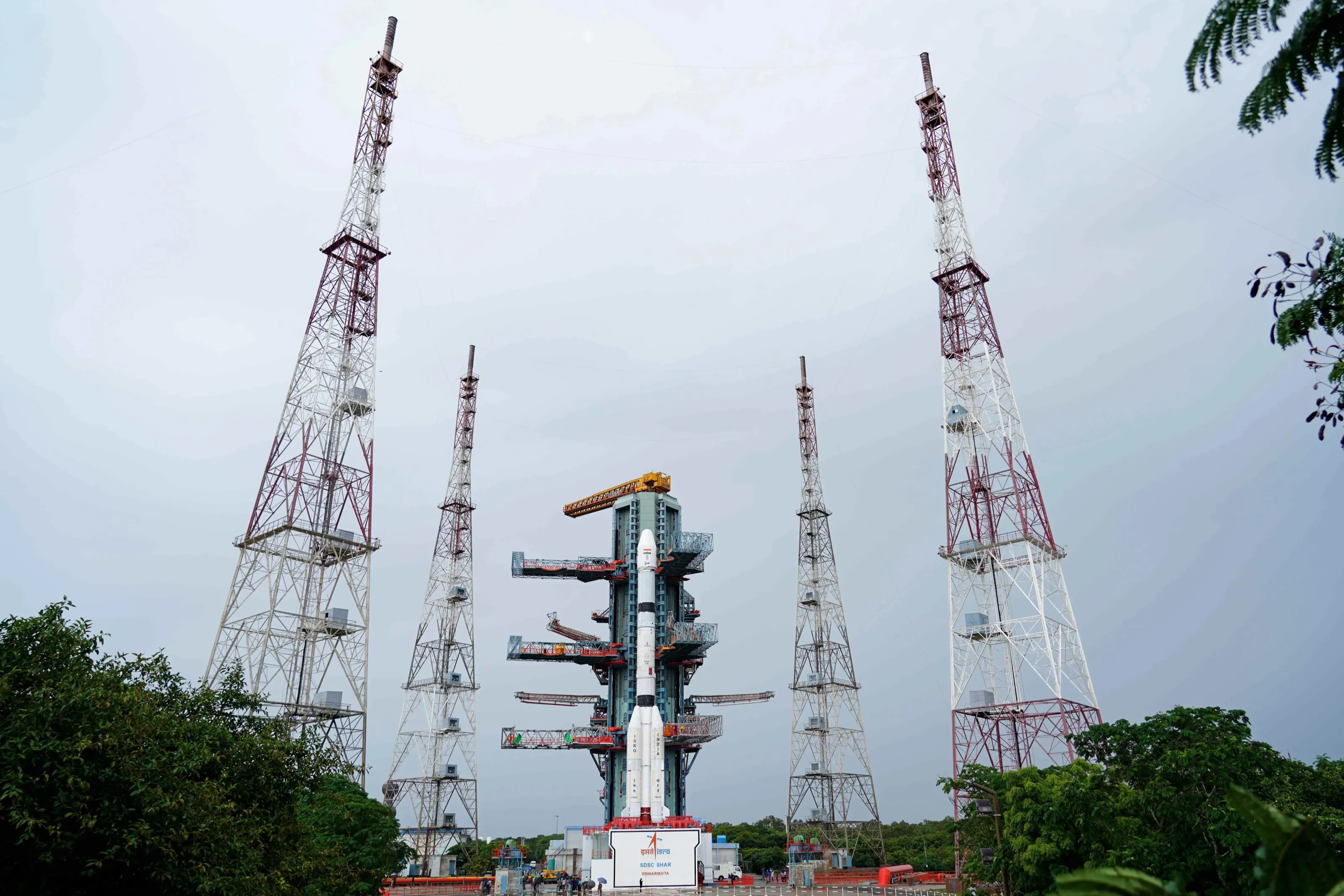 A white GSLV rocket with four strap-on boosters stands against a blue umbilical tower with grey work platforms under an orange crane, under a grey sky, between four tall red and white lattice towers, two on each side; trees frame the bottom corners and a sign at the foot of the rocket reads SDSC SHAR