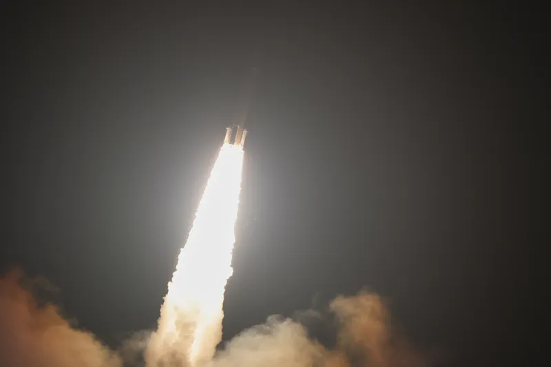 An H3 rocket climbs through a dark night sky, leaning slightly to the right: a white-yellow flame from its boosters and engines stretches down into a wide trail of lit smoke that fills the bottom of the frame, and the rocket's dark body is just visible above the glare