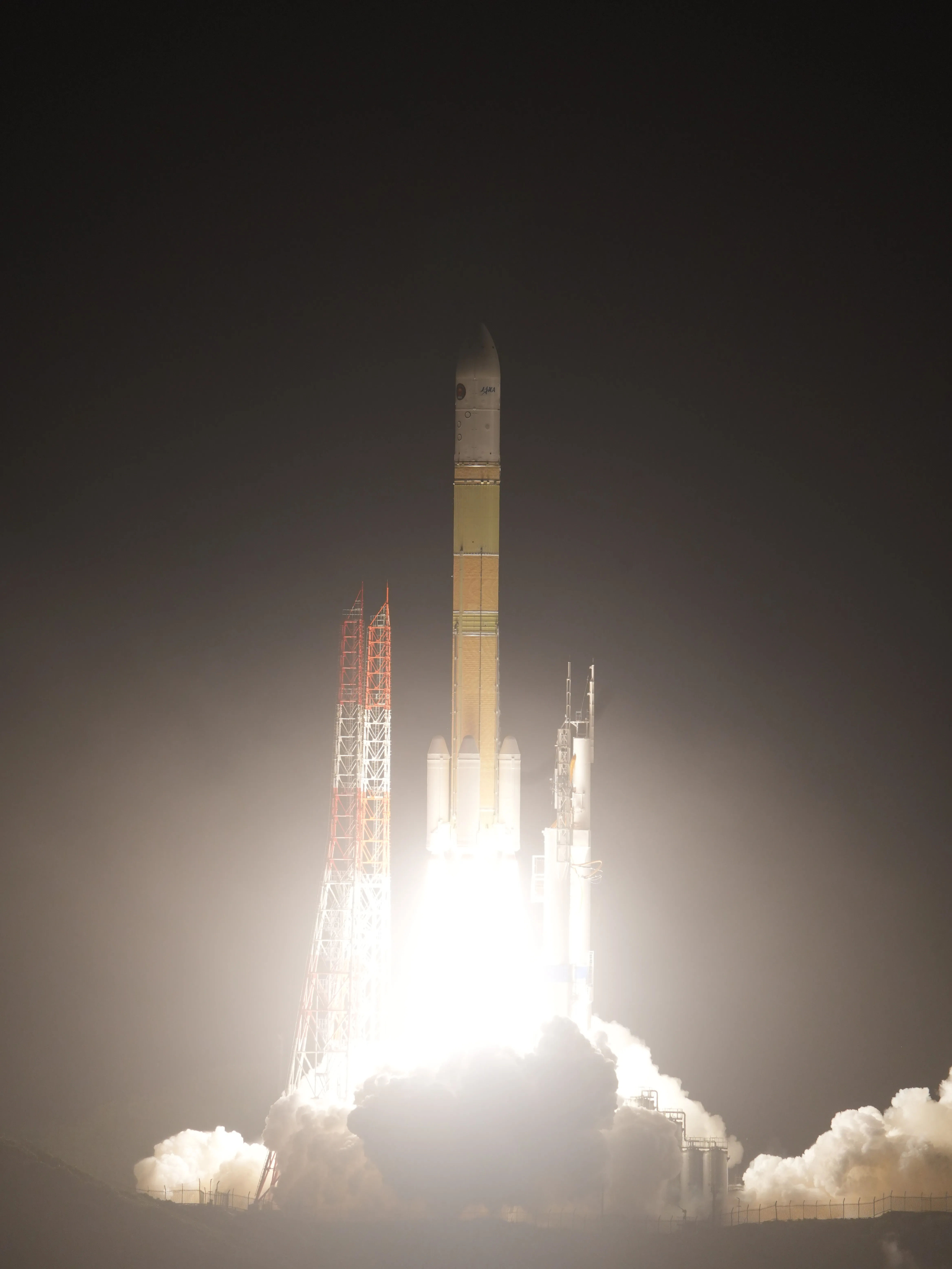 The H3 rocket rises just above its launch pad at night, lit white by its own flame: the white fairing with a JAXA logo on top, the gold-brown core stage, four white solid boosters at the base, two red-and-white lattice towers to its left, a grey umbilical tower to its right, and clouds of white exhaust rolling out below