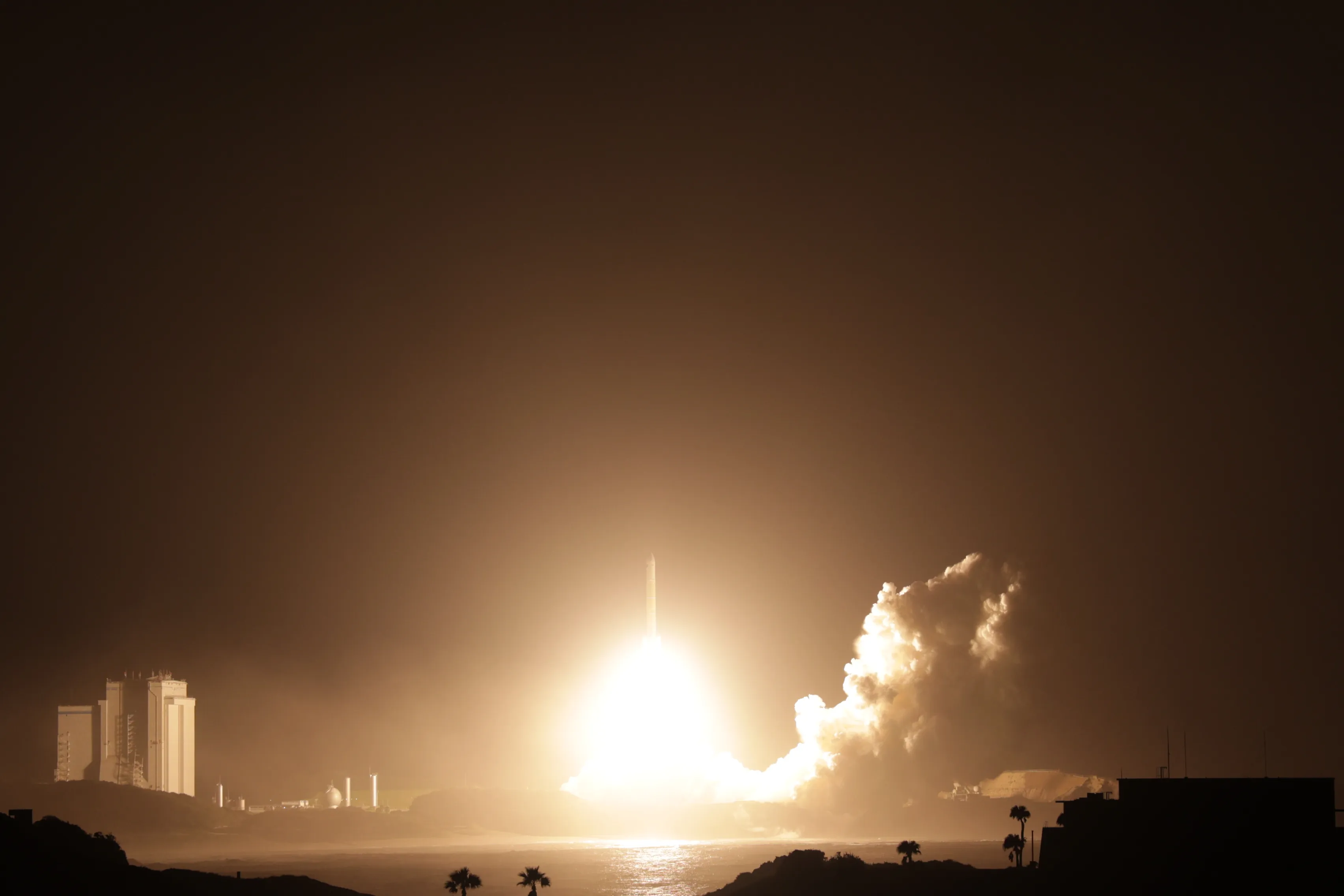 Night lift-off of an H3 rocket seen from across the water at Tanegashima: a ball of white-yellow flame at its base lights the low cloud and a wall of exhaust rolling to the right, tall white buildings stand to the left, and palm trees and a low building are silhouetted against the glowing sea in the foreground