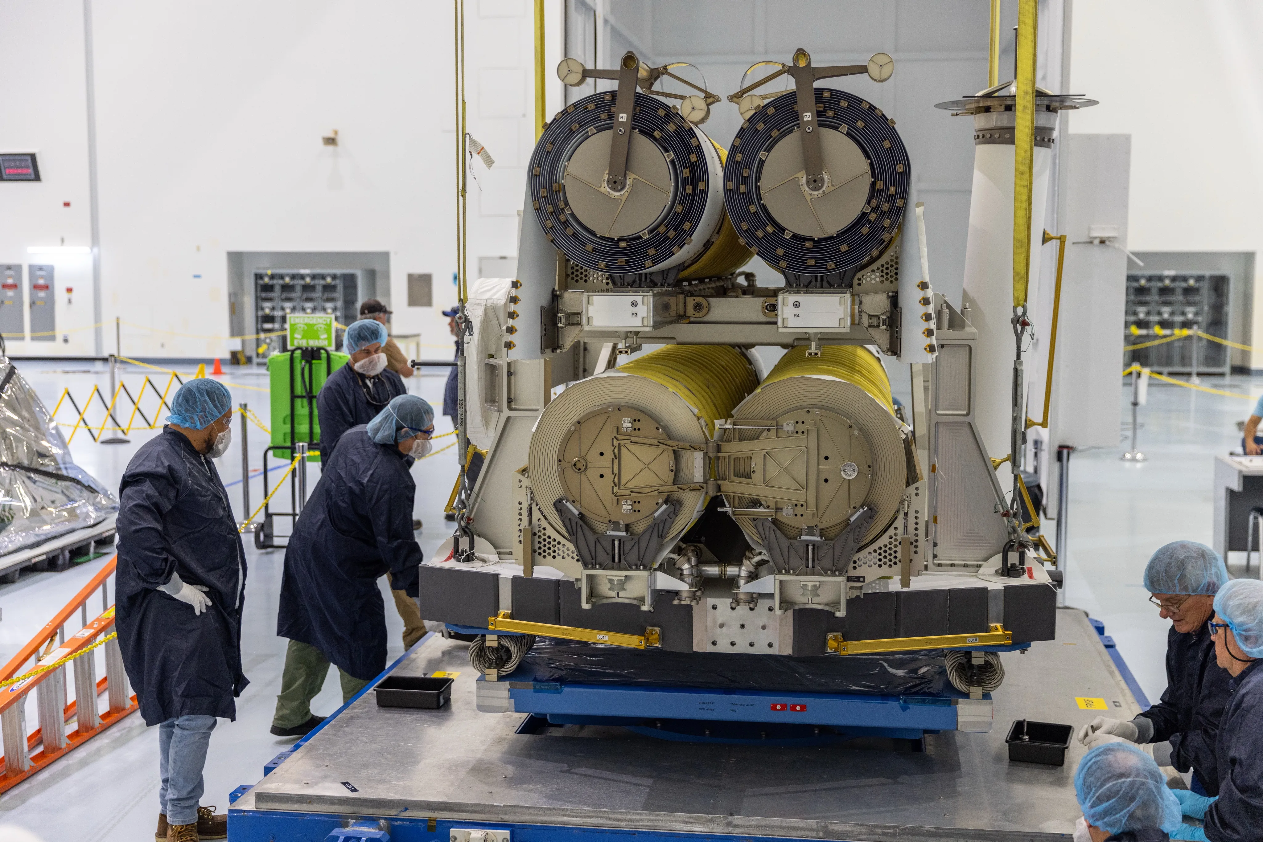 In a white clean room, a crane lowers a stacked pair of solar arrays, four large drums of rolled-up yellow blanket, the top two with dark spools on their ends, onto a blue platform, while technicians in blue gowns and hairnets guide it from both sides