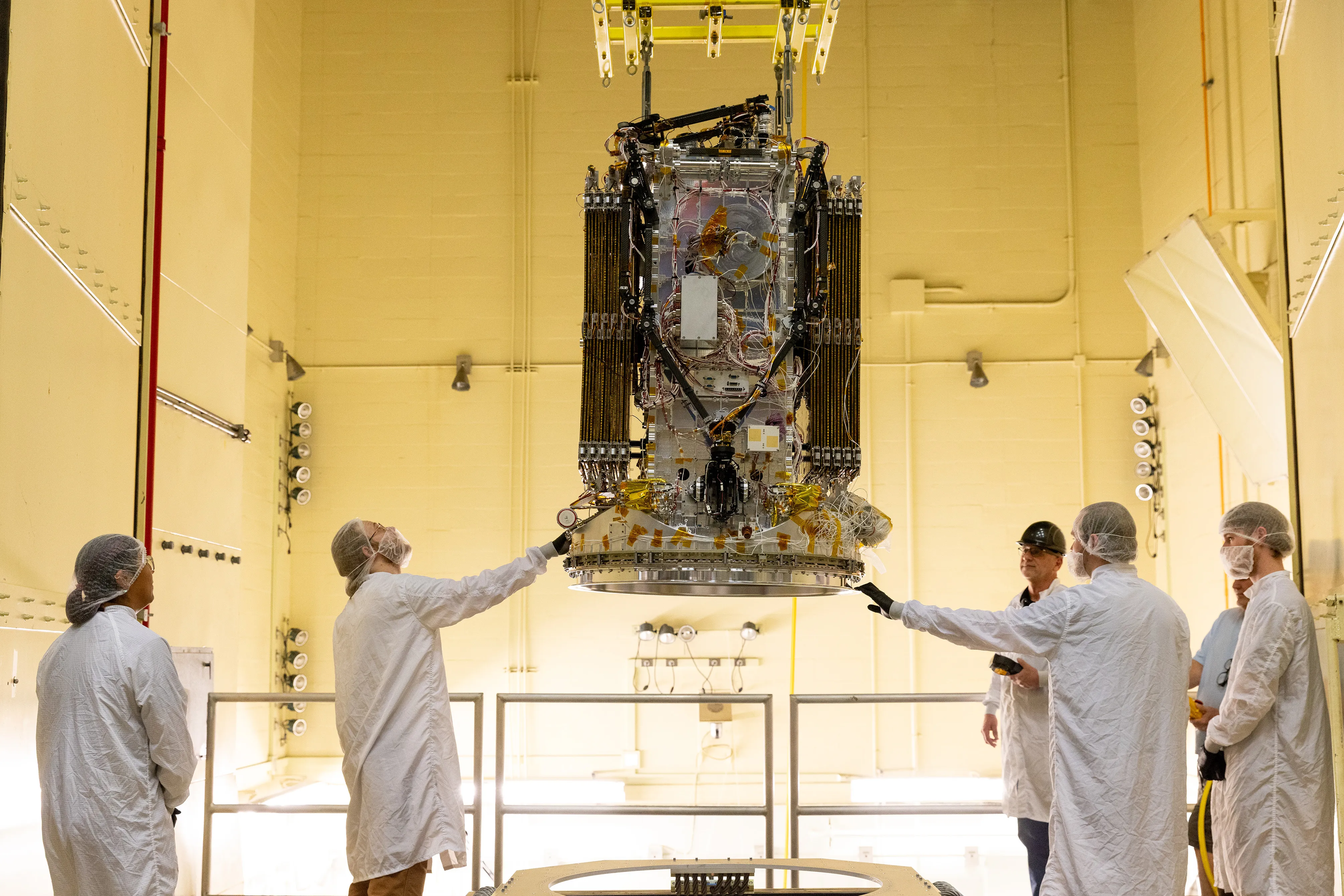 El satélite LINK, una estructura en forma de caja con los paneles solares y los brazos robóticos plegados, cuelga de una grúa sobre una mesa de vibración mientras la sujetan ingenieros con trajes de sala limpia