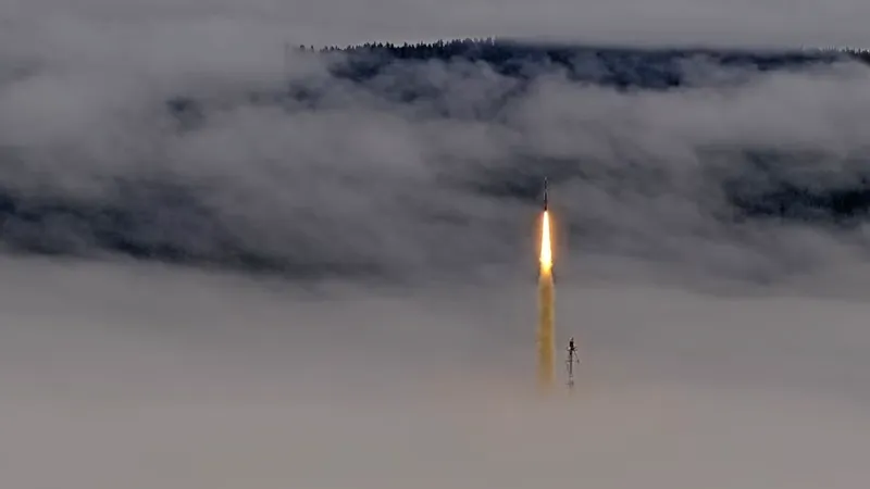 A slim dark rocket rises on a bright yellow-white flame out of a bank of low grey fog, trailing a glowing yellowish column of exhaust; just to its right the top of a thin lattice mast pokes out of the fog, and behind, a dark forested ridge shows through gaps in the cloud