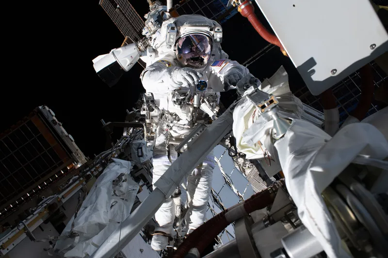 An astronaut in a white spacesuit with an American flag on the shoulder, visor down and mirroring the station, holds a tool at his chest and a rail with his other hand, among white covers, red hoses and dark solar panels, with a white capsule docked beyond him against black space