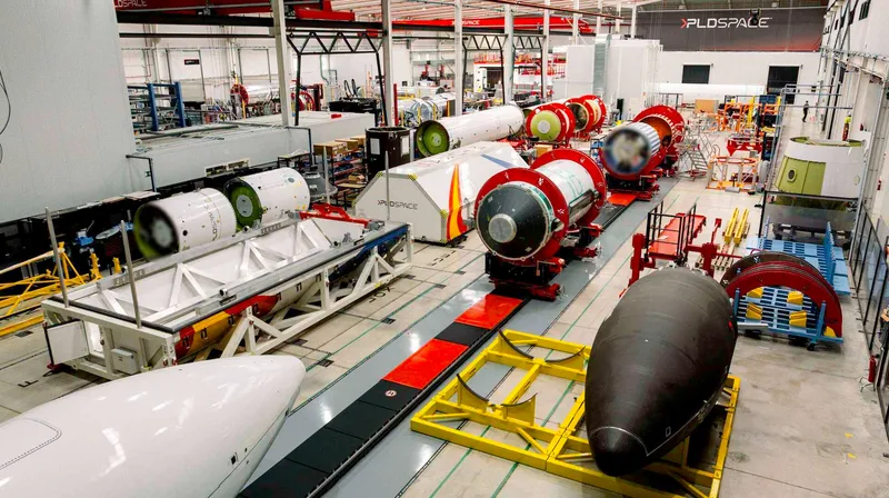 A long factory hall seen from above: white and black rocket stages and tank sections with orange end rings rest on cradles in rows along a grey floor, a white fairing half lies in the foreground and a black one on a yellow stand to the right, and the PLD Space logo is on the far wall