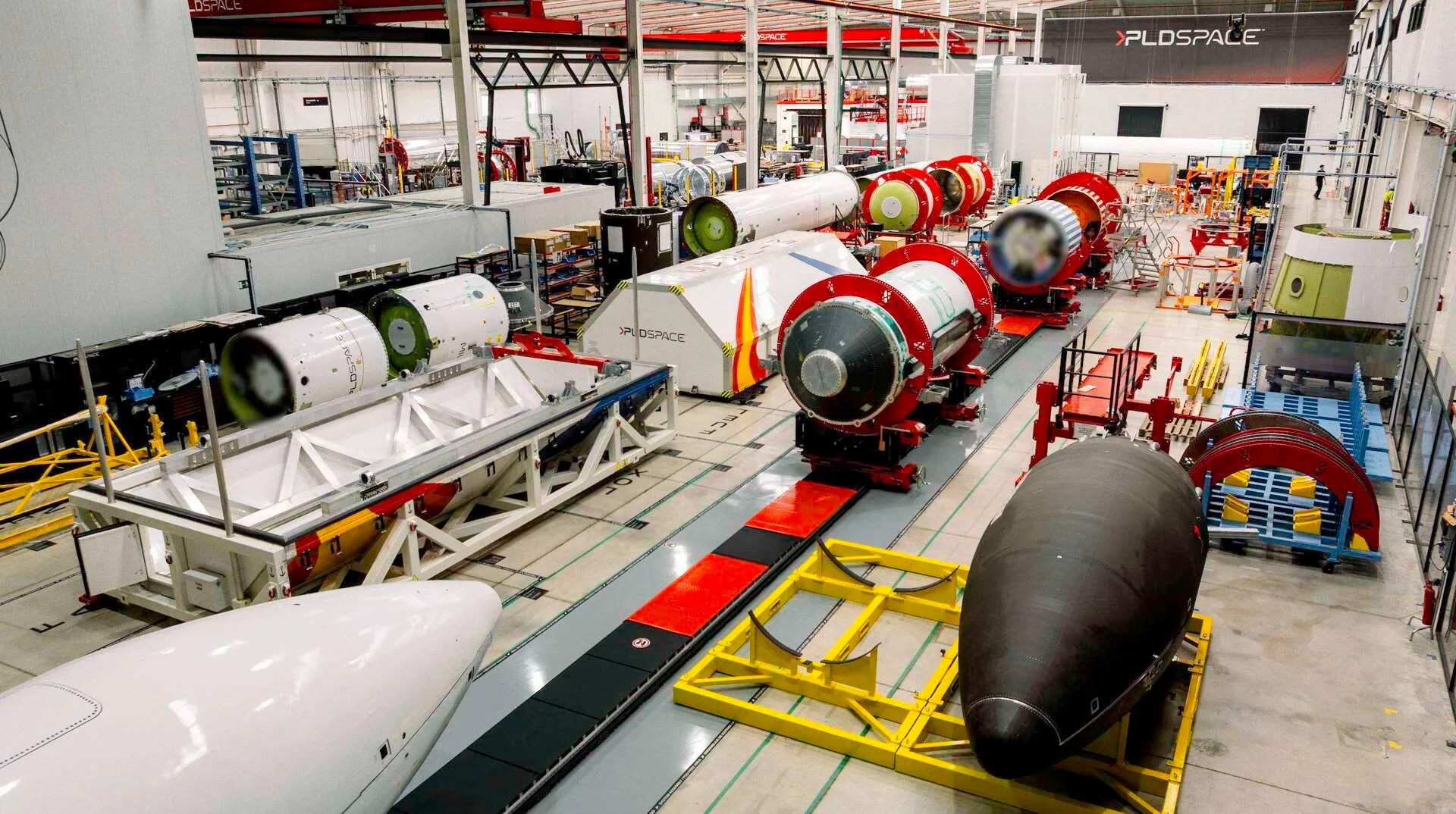A long factory hall seen from above: white and black rocket stages and tank sections with orange end rings rest on cradles in rows along a grey floor, a white fairing half lies in the foreground and a black one on a yellow stand to the right, and the PLD Space logo is on the far wall