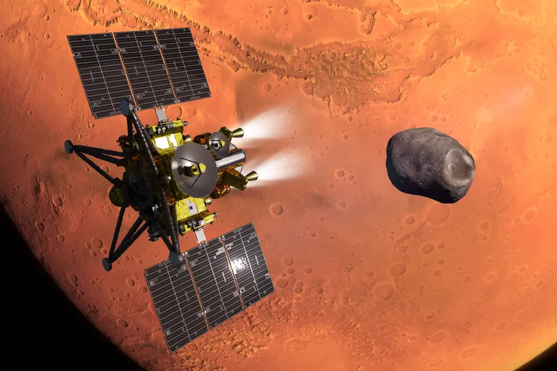 JAXA's rendering of the MMX spacecraft over the orange, cratered face of Mars: a box wrapped in gold foil, with two dark solar wings, a round white dish antenna and four long black landing legs, fires two thrusters that glow white, while the small, lumpy grey moon Phobos hangs to its right