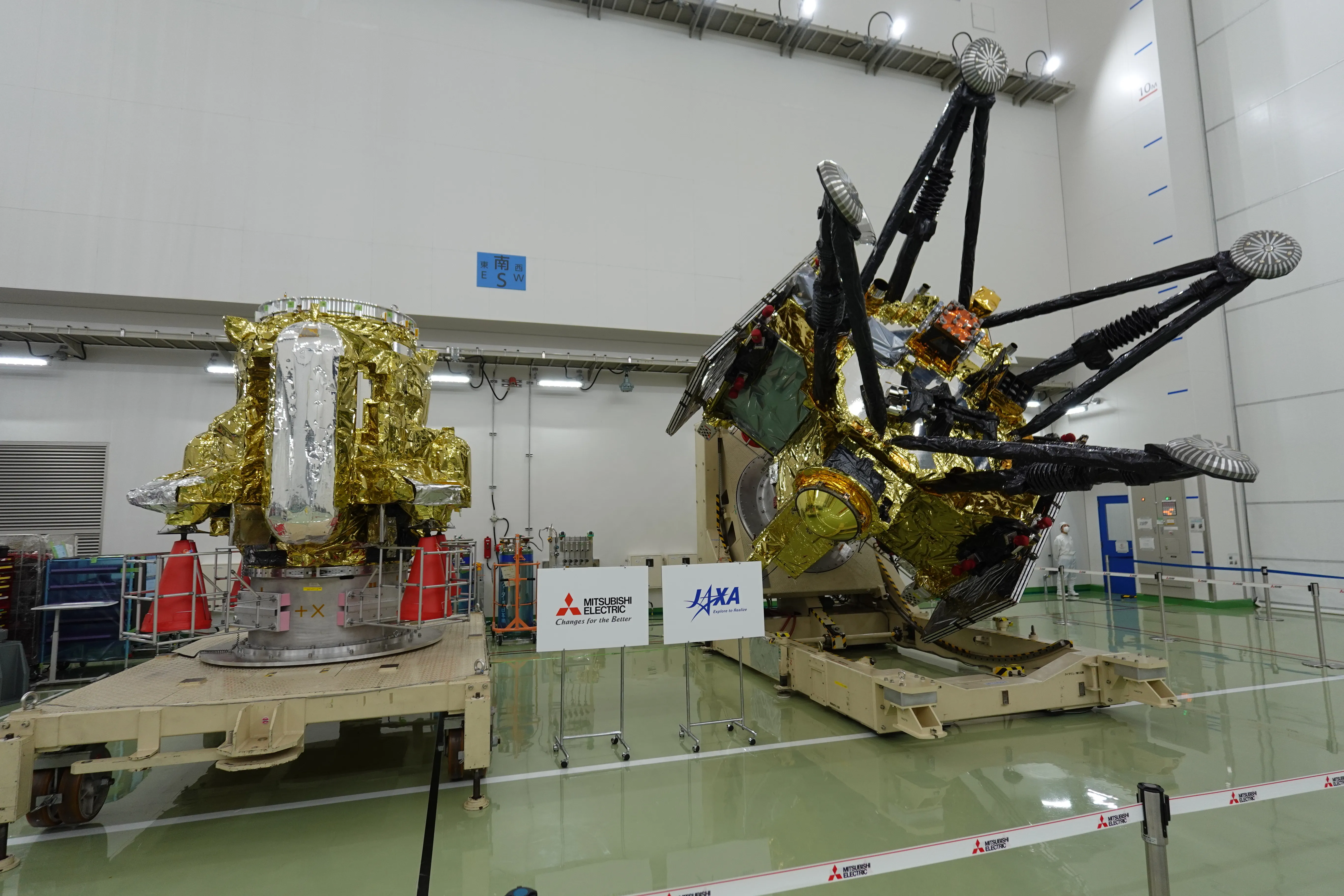 In a large white hall with a green floor, the two parts of the MMX spacecraft sit on cream-coloured transport stands: on the left a squat module wrapped in gold foil with a silver-wrapped cylinder down its front, on the right a bigger gold module tilted on its side with four long black landing legs ending in round footpads, and between them two signs reading Mitsubishi Electric and JAXA