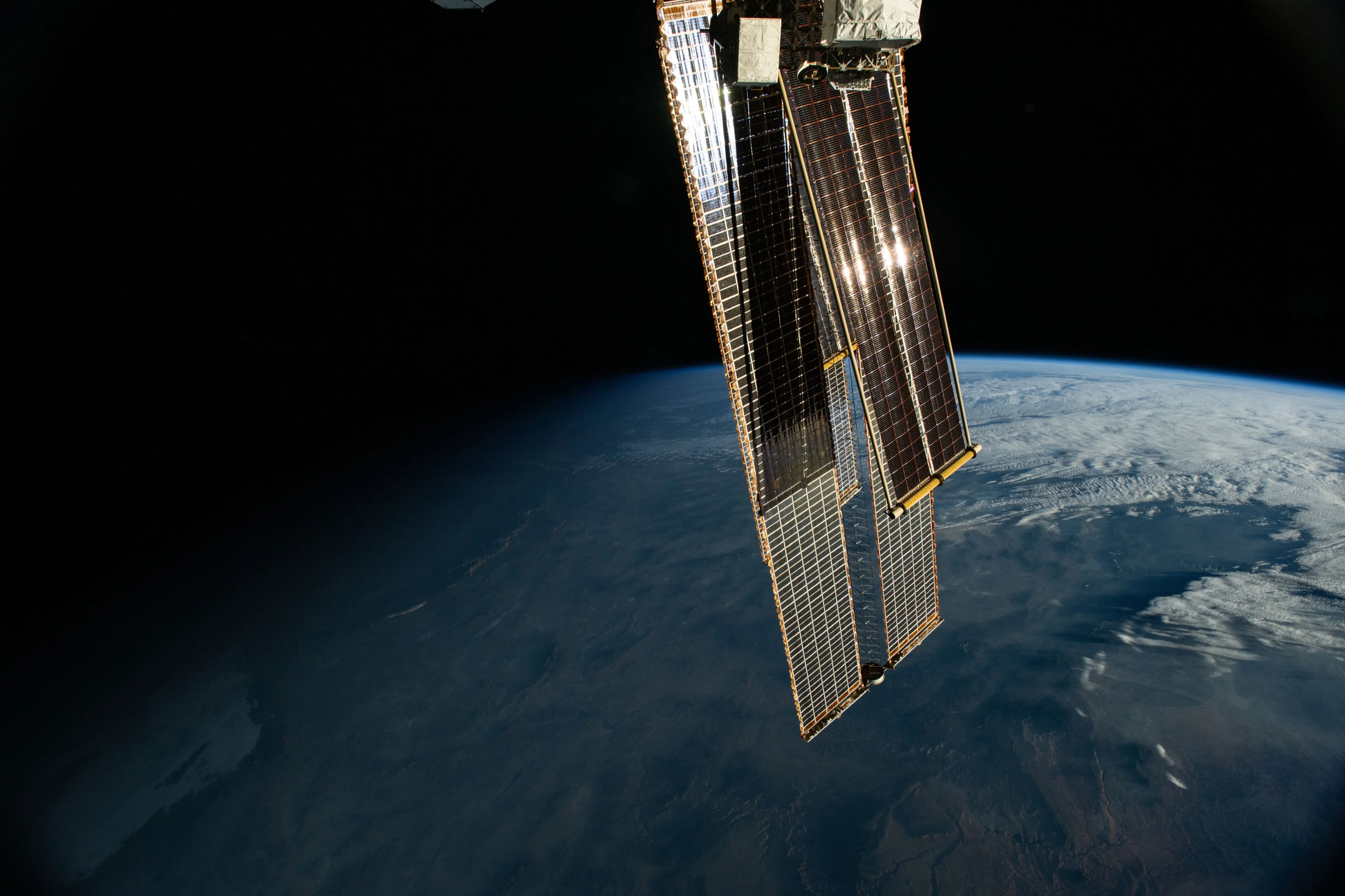 A roll-out solar array, laid in front of one of the station's original arrays, catches the first sunlight on its dark cells against black space, above the curve of the Earth, dim blue and brown below with white cloud to the right
