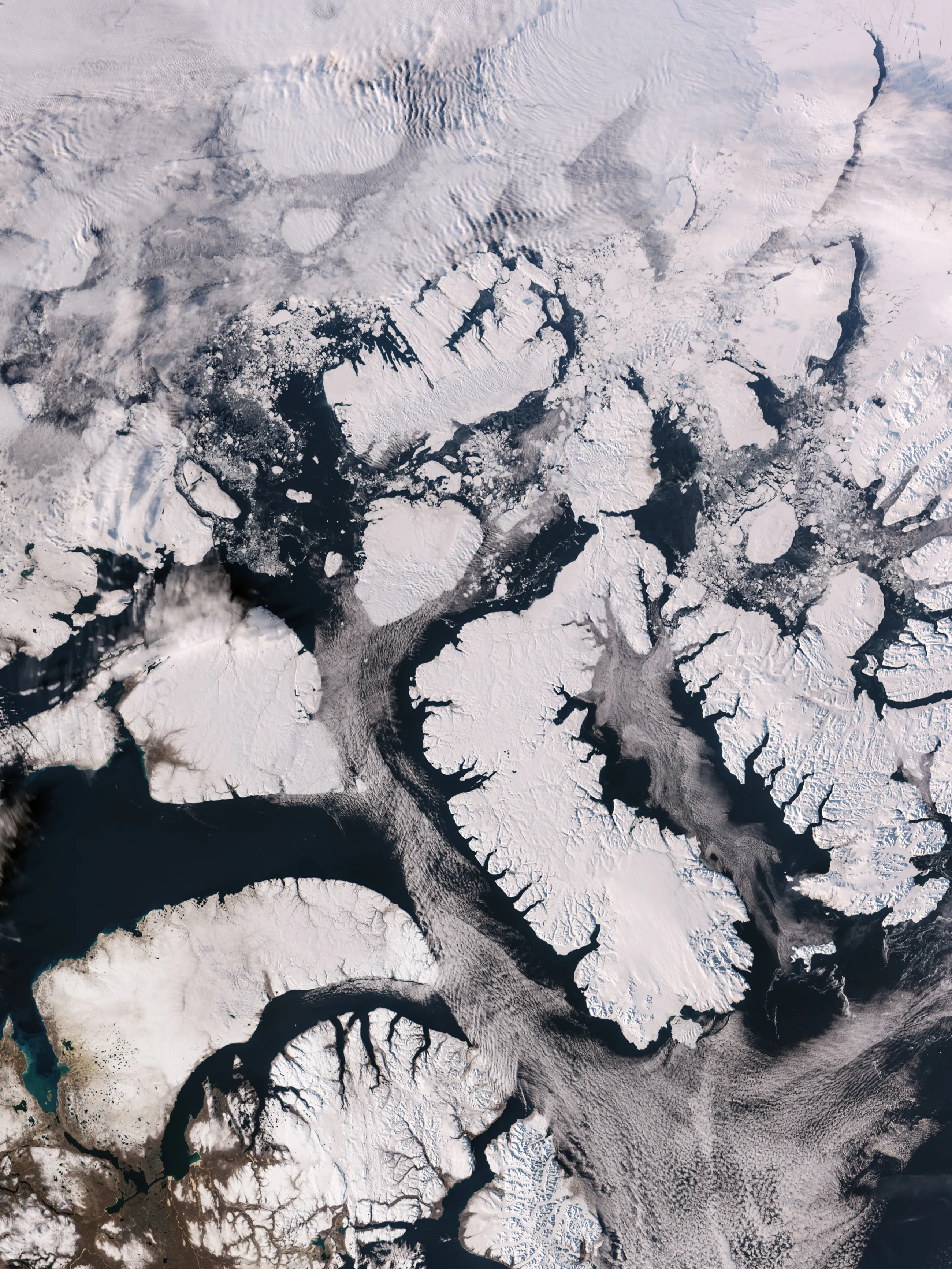 Snow-white islands cut by dark, branching fjords, surrounded by dark open water with scattered broken ice, thick, rippled white cloud across the top third and a patch of brown, snow-free land at the lower left