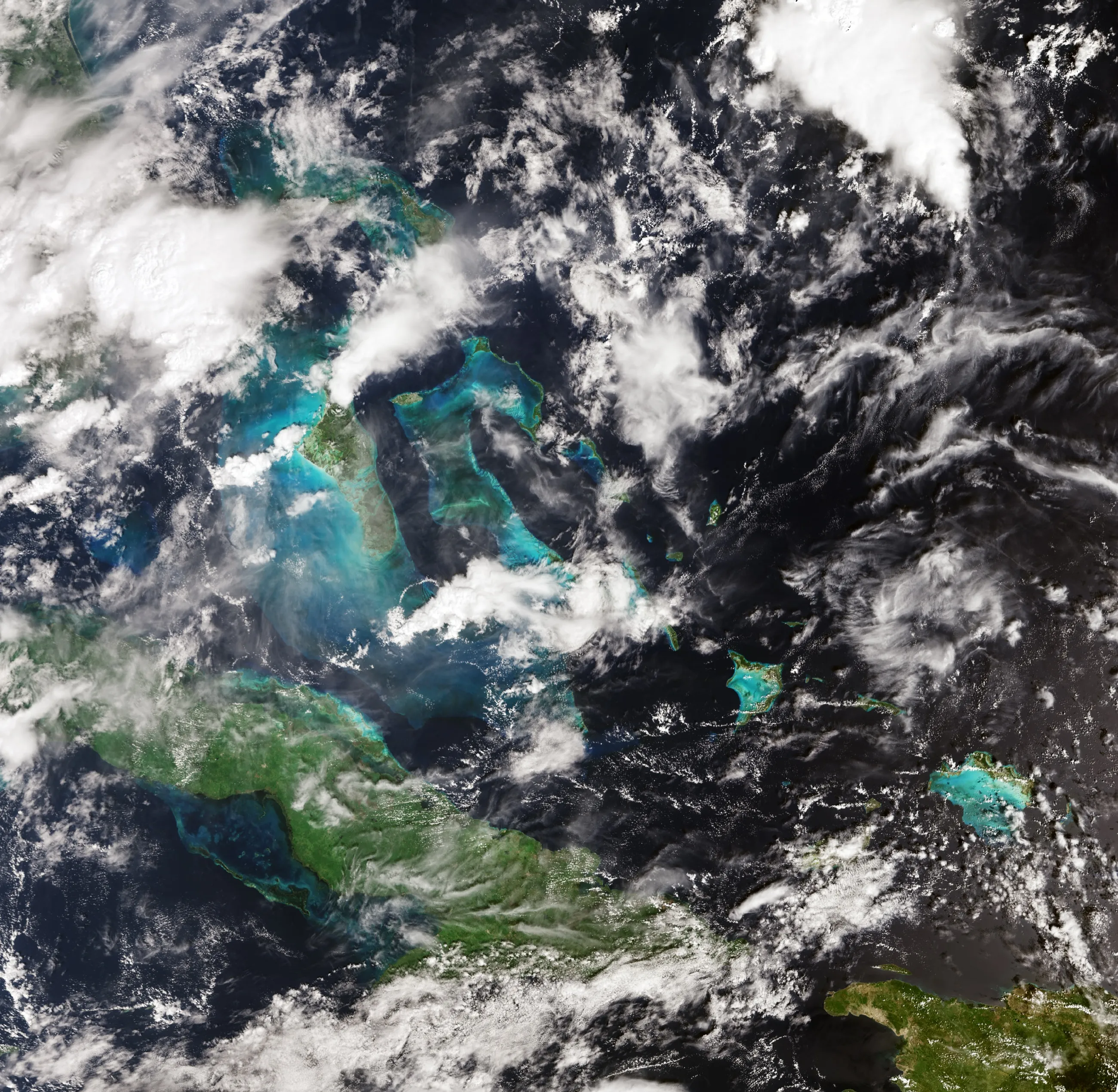 Green islands set in patches of turquoise shallow water in a dark blue sea, scattered with white cloud; a long green island stretches across the lower part of the picture