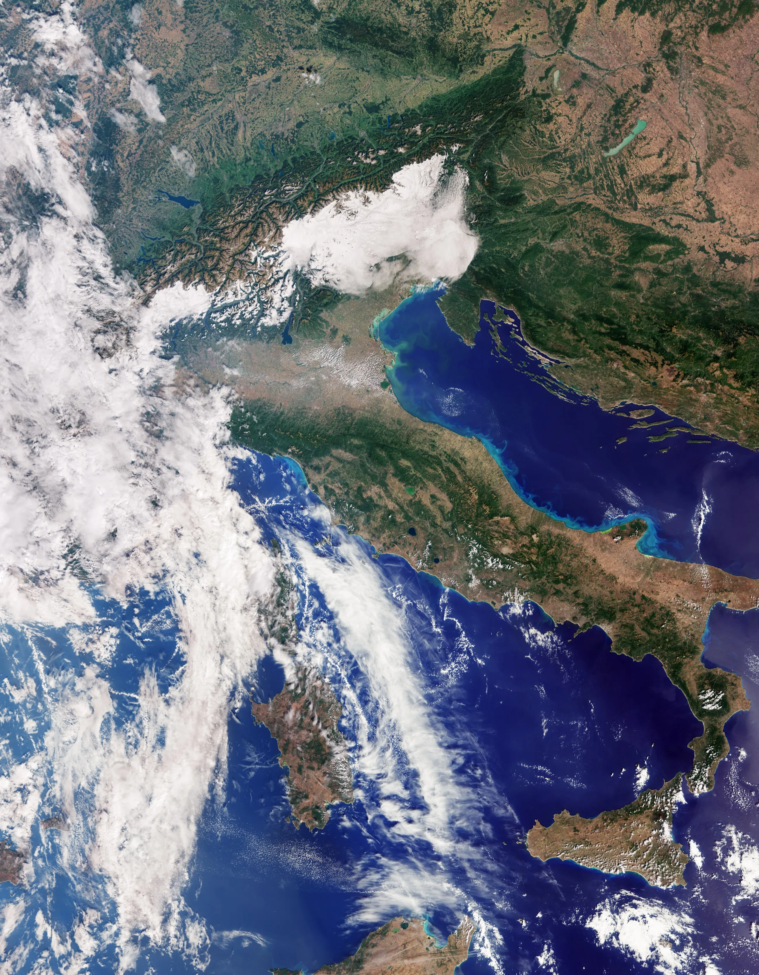 Italy from orbit in natural colour, from the green plains and the Alps at the top down to Sicily near the bottom; a long band of white cloud covers the left of the picture, Sardinia shows through it in the deep blue sea, and pale turquoise water lines the Adriatic coast