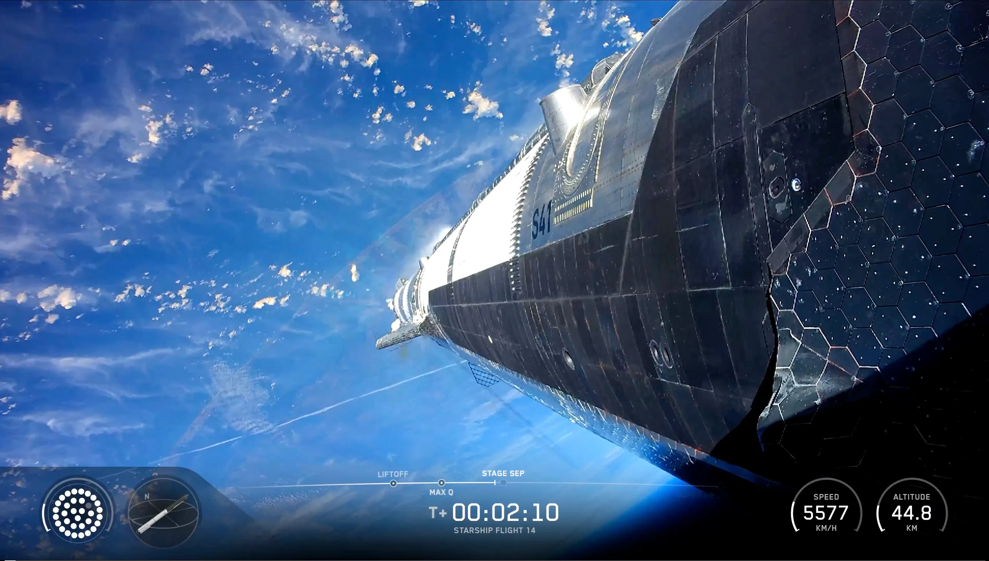 Ship 41 seen from a camera on its own hull as it climbs, still attached to the Super Heavy booster: black heat-shield tiles and the letters S41 on the steel in the foreground, blue ocean and scattered white clouds far below. The webcast readout reads T+00:02:10, speed 5577 km/h, altitude 44.8 km