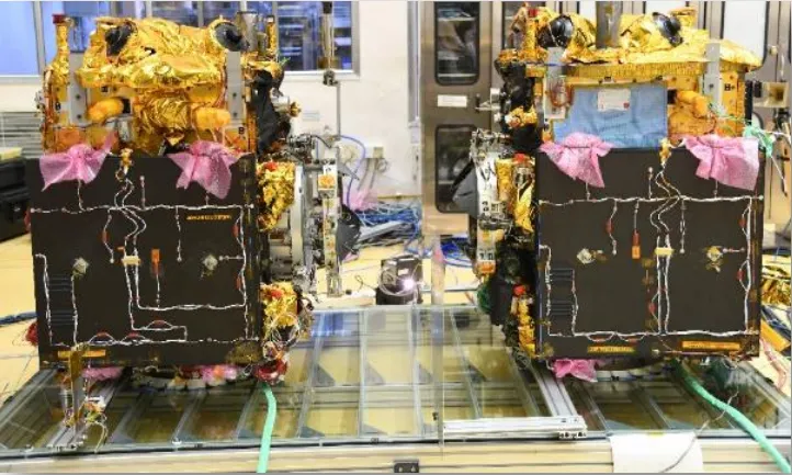 Two boxy satellites stand side by side on a glass-topped test frame in a laboratory. Each is wrapped in crinkled gold foil on top, with a large black side panel laced with thin white cables and dotted with small fittings; pink protective covers hang from the top corners, and cables trail across the floor between them