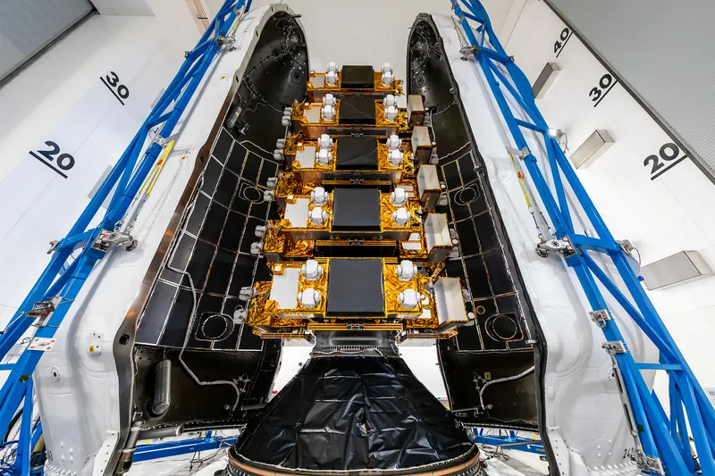 A stack of flat satellites wrapped in gold foil stands between the two open halves of a rocket fairing, white outside and black inside; the top of each satellite carries a large black square panel and four white turret-like units, a cone wrapped in black film holds the stack at the bottom, and blue access frames flank the fairing in a white hall whose walls are marked 20, 30 and 40