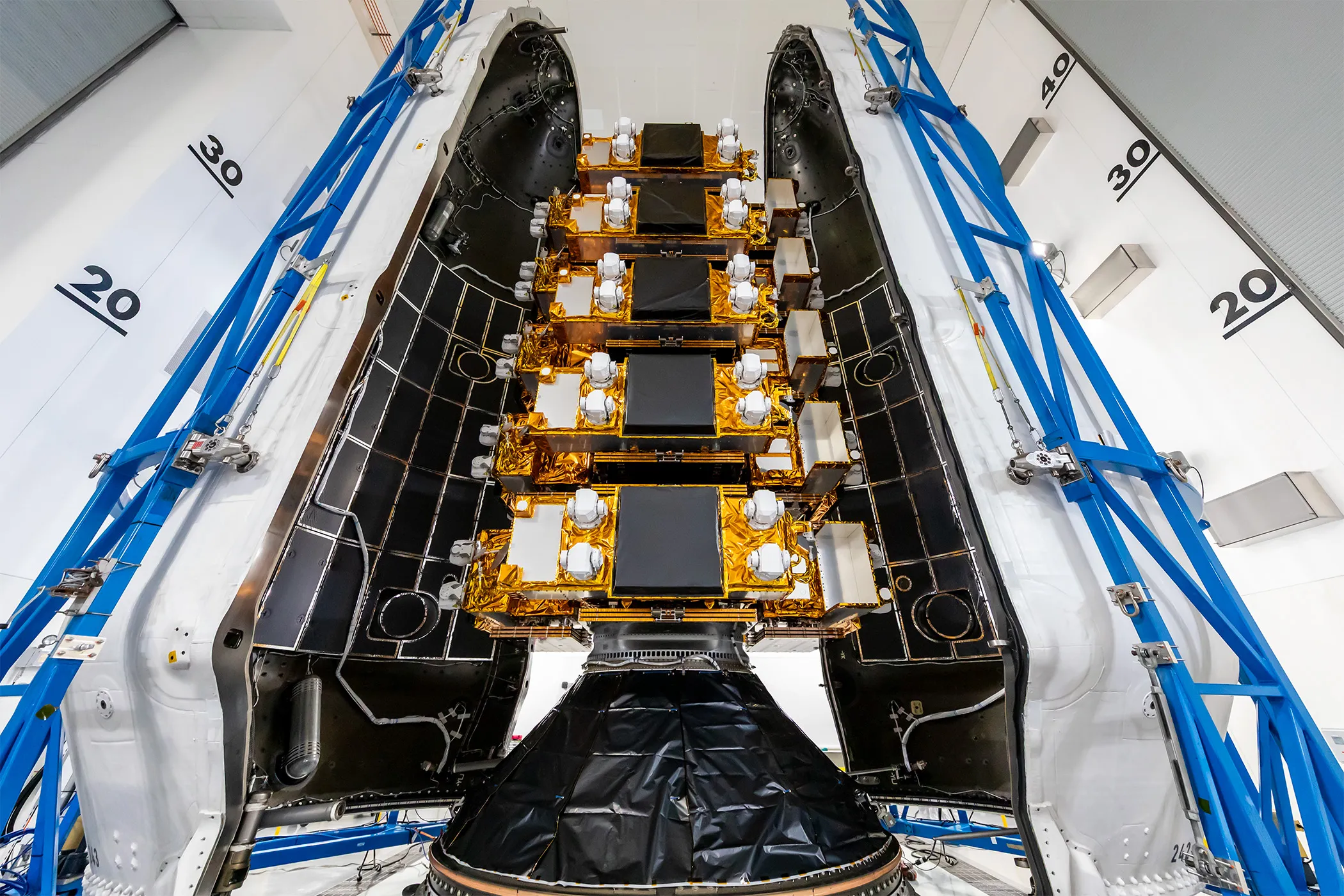 A stack of flat satellites wrapped in gold foil stands between the two open halves of a rocket fairing, white outside and black inside; the top of each satellite carries a large black square panel and four white turret-like units, a cone wrapped in black film holds the stack at the bottom, and blue access frames flank the fairing in a white hall whose walls are marked 20, 30 and 40