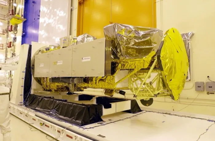 The Aqua satellite in gold and silver thermal blankets, the round dish of an antenna at one end, lying on its stand in a clean room at Vandenberg before its launch in 2002.
