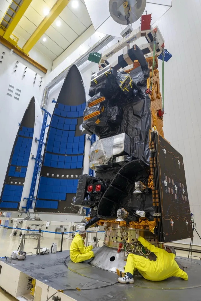 MetOp-SG A1 standing on its Ariane 6 launch adaptor in Kourou in August 2025, dark thermal blankets over its instruments, the two halves of the rocket’s fairing behind it and two technicians at its base.