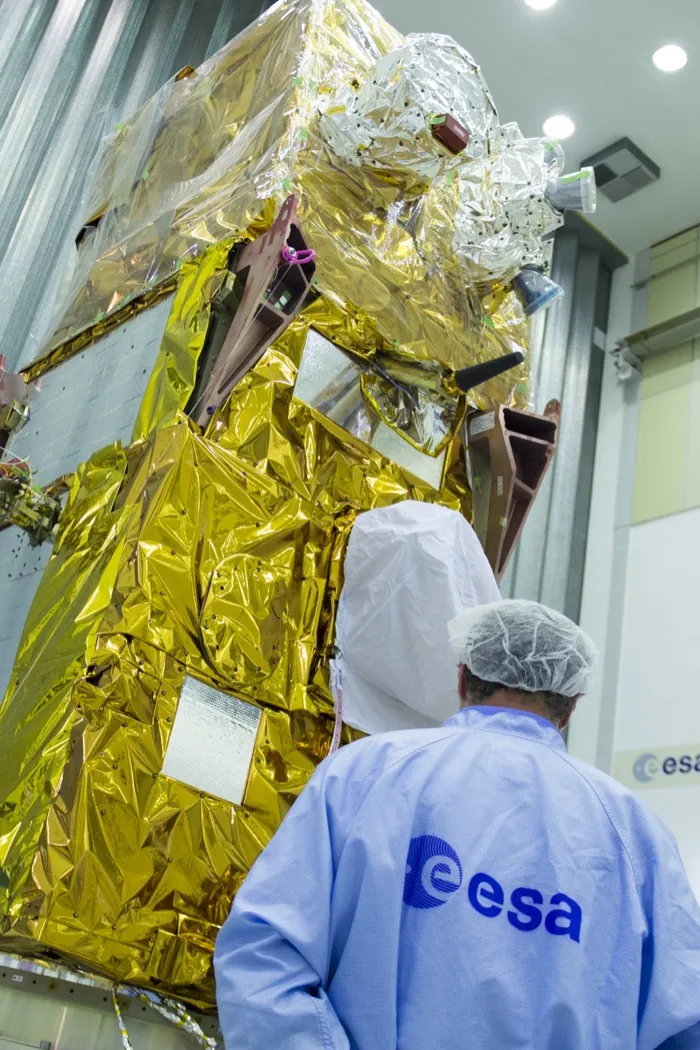 The Sentinel-2B satellite wrapped in gold insulation at ESA’s test centre in the Netherlands in 2016, an engineer in an ESA smock in front of it.