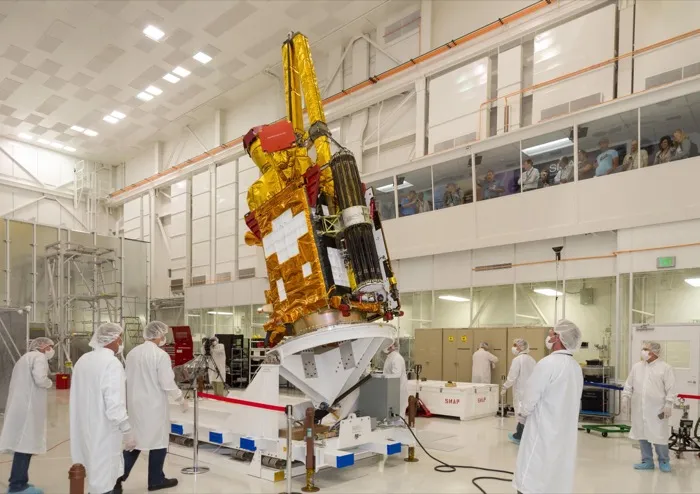 The SMAP satellite in gold insulation, its antenna boom and folded mesh reflector on top, lowered onto a stand in a clean room at NASA’s Jet Propulsion Laboratory in 2014, engineers in white suits around it.