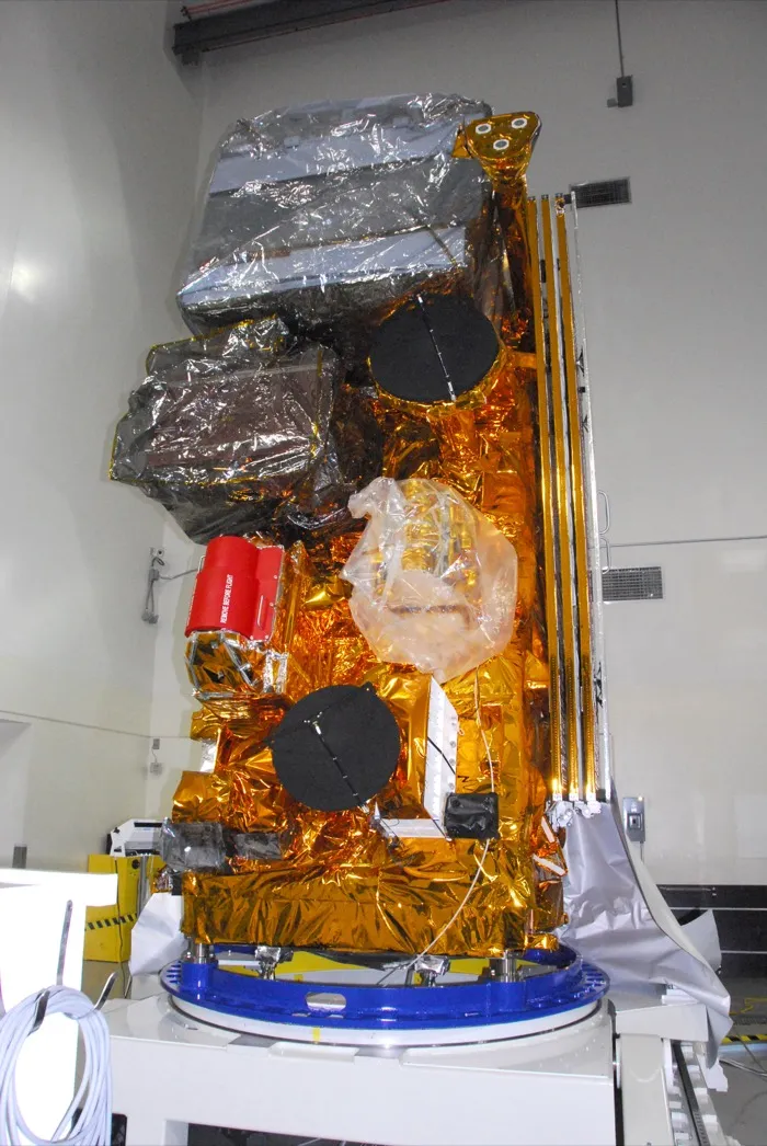 The Suomi NPP satellite standing upright in a clean room at Vandenberg before its launch in 2011, wrapped in gold and silver insulation, its solar array folded along one side.