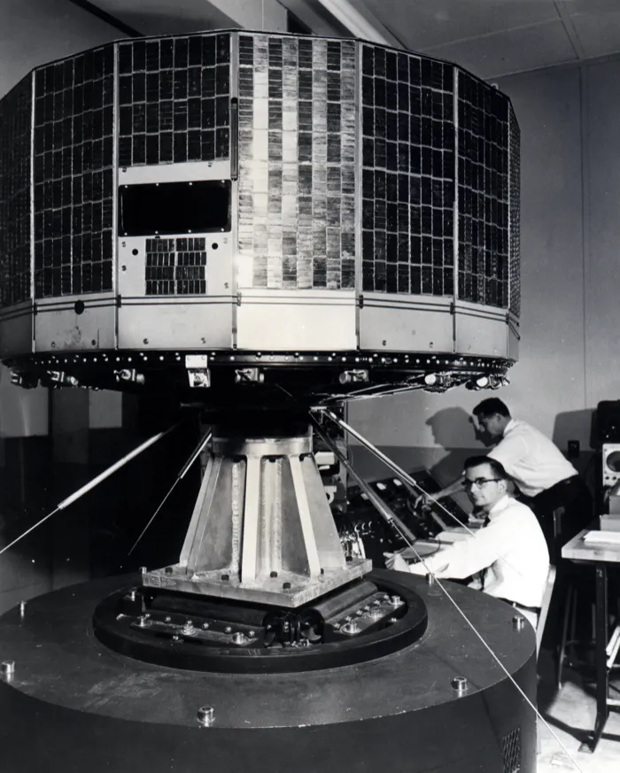 TIROS-1, an 18-sided drum tiled with solar cells, on a vibration test stand at RCA in Princeton in 1960, two engineers at the controls behind it.
